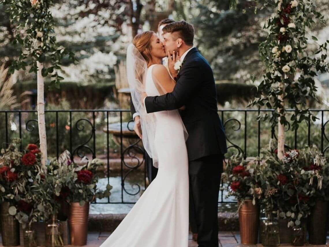 Amidst a stunning fall wedding, the bride and groom share a kiss during their outdoor ceremony. They are surrounded by floral arrangements with red and white flowers and lush greenery, while the background features trees and a wrought iron railing, perfectly capturing the season's charm.