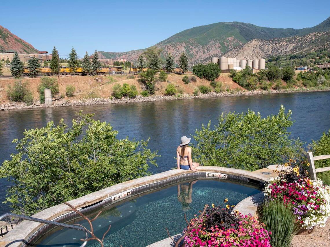 A person in swimwear and a hat relaxes by the edge of a curved pool overlooking a scenic river, ready for various activities. Colorful flowers bloom nearby, and lush mountains rise in the background under a clear blue sky.