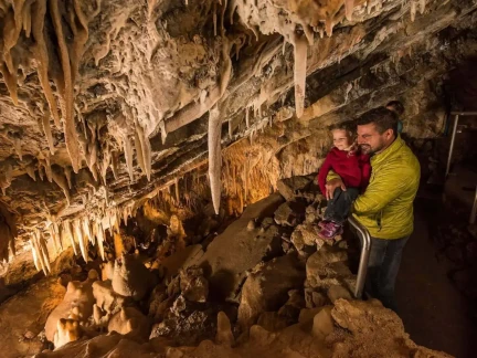 A father holding his child stands inside an adventure-filled cave adorned with numerous stalactites and stalagmites. The scene is warmly lit, highlighting the intricate rock formations on the ceiling and floor, inviting exploration of nature's wonders.