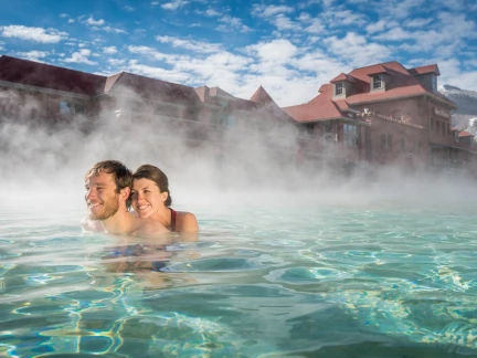 A couple enjoys swimming in a steaming outdoor pool under a blue sky with scattered clouds, as gentle splashes ripple around them. In the background, a red-roofed building is partially visible.