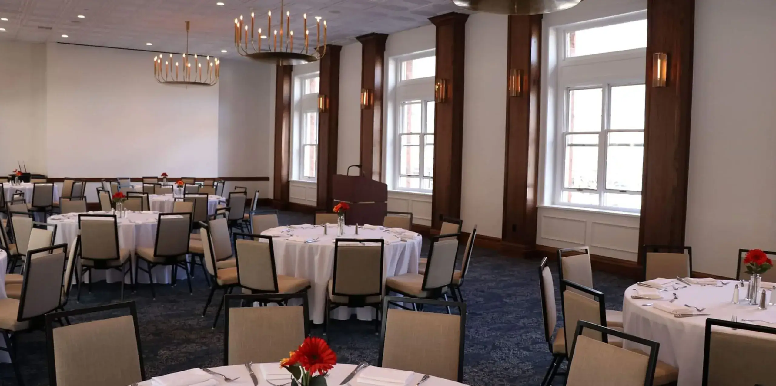 A corporate banquet room features round tables draped in white tablecloths, each adorned with a red flower centerpiece. Beige chairs surround the tables, while large windows line the wall and elegant chandeliers hang from the ceiling.