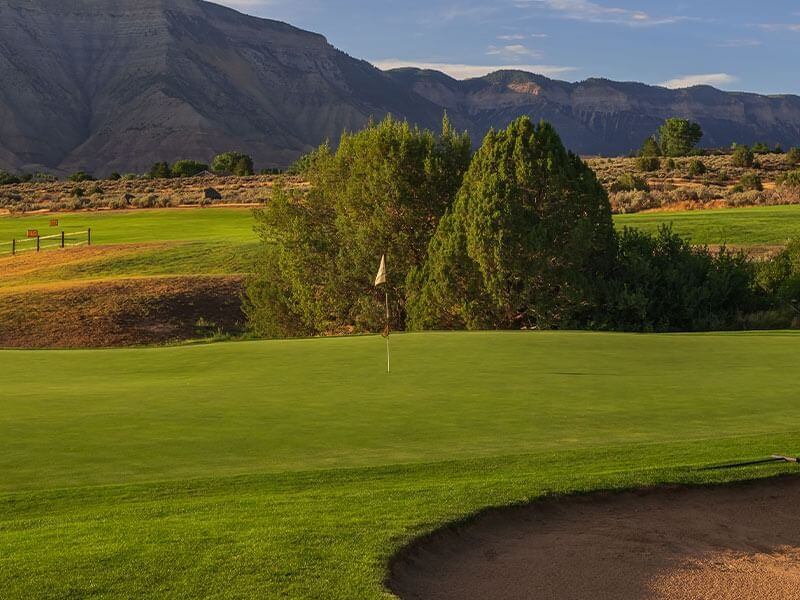 A lush green golf course with a flagstick on the putting green, perfect for any golf enthusiast, surrounded by trees and shrubs. In the background, a range of rocky mountains under a partly cloudy blue sky. A sand bunker is visible in the foreground.