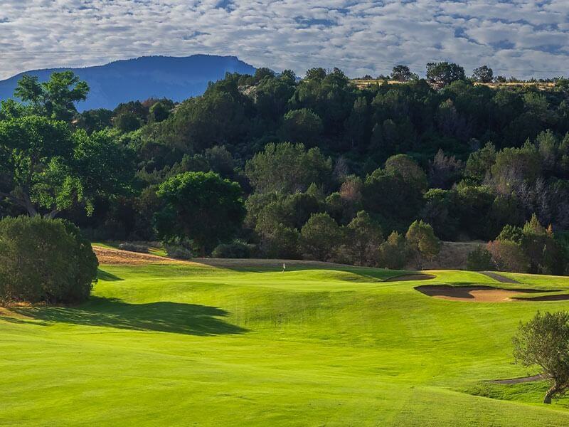 A picturesque golf course featuring lush, green fairways and a challenging sand trap under a partly cloudy sky. The course is surrounded by dense trees, with the silhouette of a mountain in the background—a golfer's paradise.