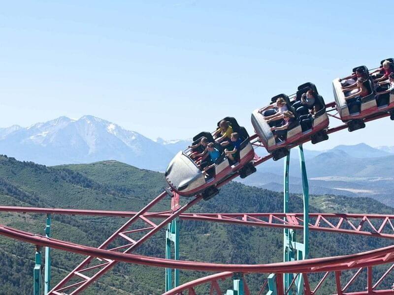 A roller coaster with several people riding twists along red tracks against a backdrop of green hills and snow-capped mountains under a clear blue sky, offering an exhilarating spring break experience.