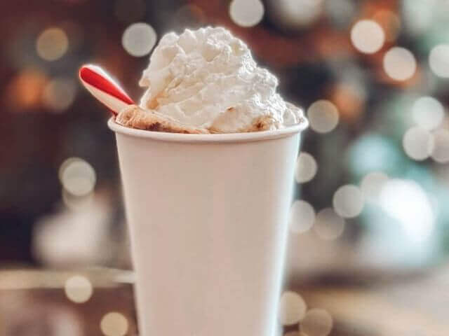 A white paper cup filled with a drink topped with whipped cream and a red straw, reminiscent of a cozy shop ambiance, is set against a blurred background with circular bokeh lights.