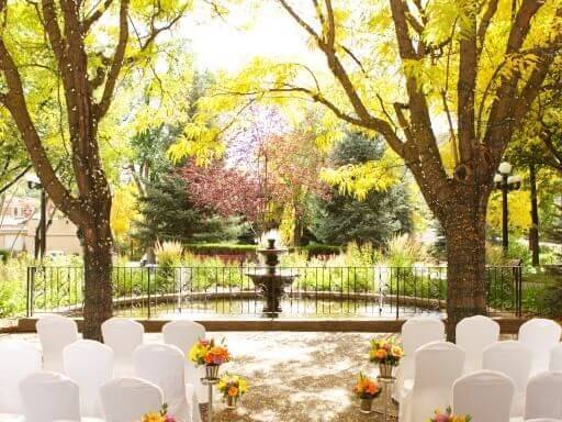 A picturesque fall wedding venue with white chairs arranged in rows, facing a black fountain surrounded by lush greenery and colorful autumn trees. Bright yellow leaves frame the scene, creating a warm and inviting atmosphere.