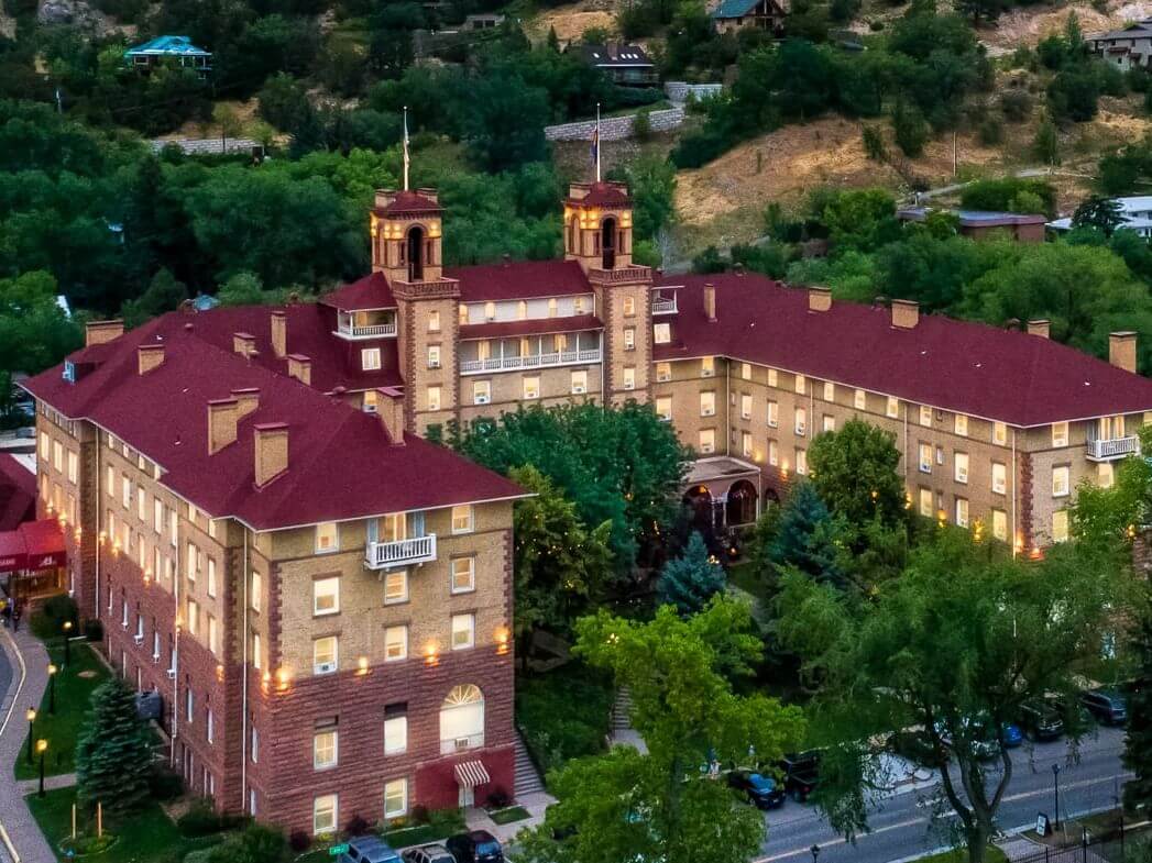 An aerial view captures the historic hotel, perfect for a spring break getaway, with its red roof and brick walls nestled among lush trees and hills. Majestic towers rise as numerous windows glow warmly from within.