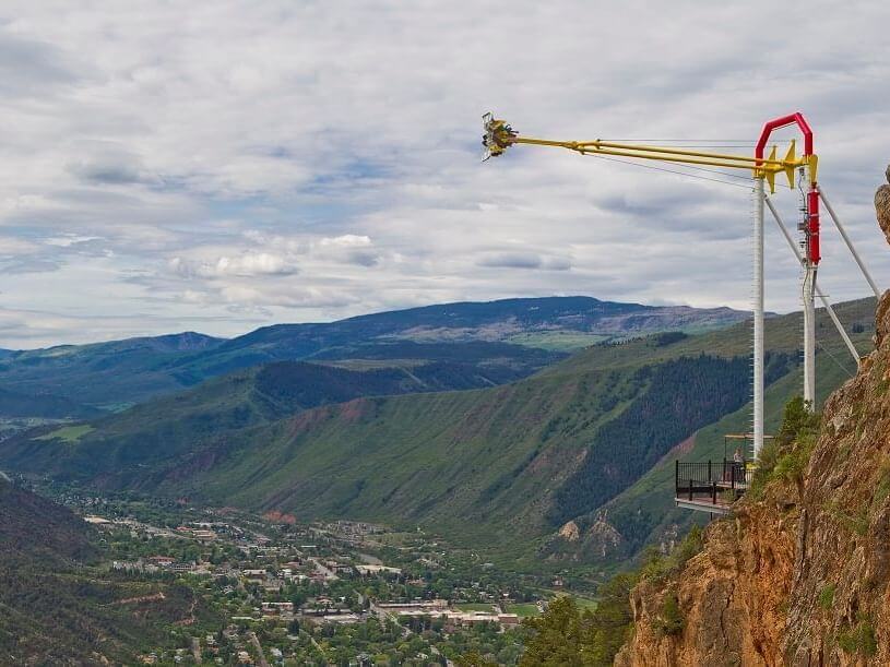 Amusement ride extending over a cliff, with people in seats swinging outward over a valley. It's the perfect spring break adventure; below, a small town is nestled among green hills, and a cloudy sky stretches above.