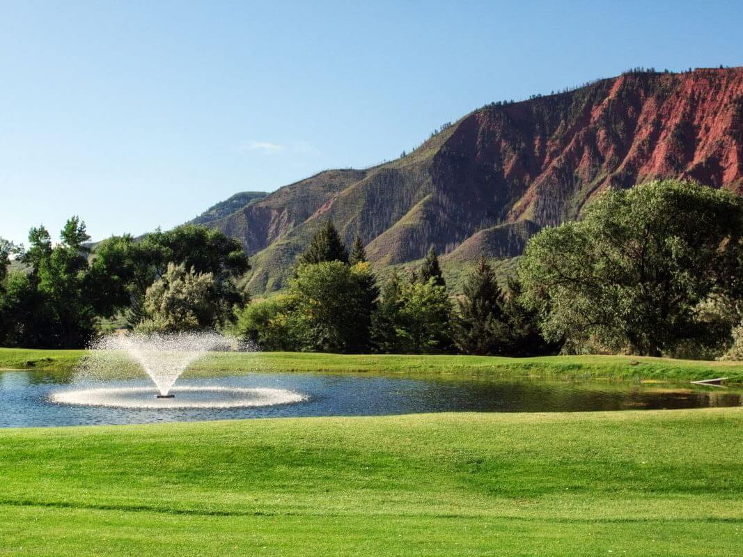 A picturesque landscape featuring a green field reminiscent of a golf course, with a water fountain in the foreground, surrounded by lush trees. In the background, red-hued mountains rise against a clear blue sky.