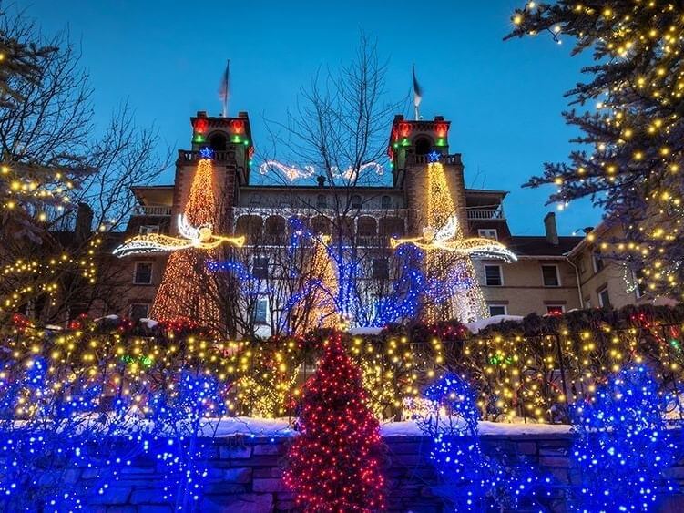 A historic building adorned with vibrant holiday lights, featuring red, blue, and gold decorations, invites visitors to indulge in holiday shopping. Two large, lit angel figures stand prominently in front, as snow blankets the ground and surrounding trees against a twilight sky.