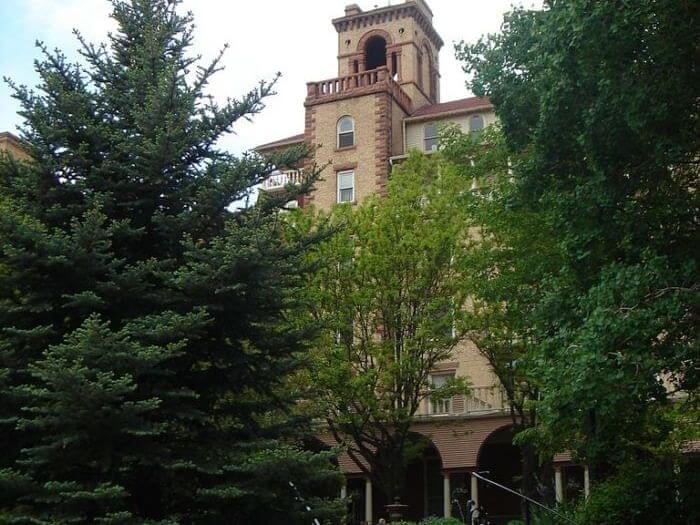 A brick building with a tall tower peeks through lush green trees, reminiscent of views on an Amtrak ride. Arched windows and balconies are partially obscured by foliage, while a clear sky provides a bright backdrop to the scene.