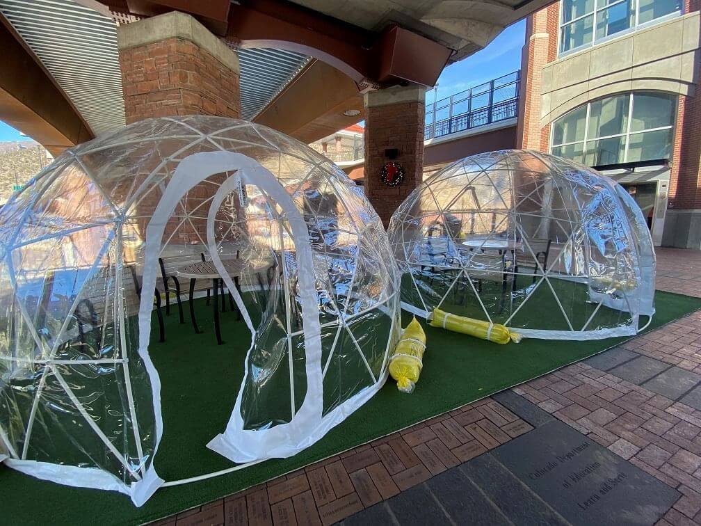 Two transparent dome structures are set up outdoors on artificial grass near brick buildings. The domes contain tables and chairs, offering a sheltered seating area. Rolled-up yellow tarps lie beside them, adding a hint of versatility to this inviting outdoor setting.