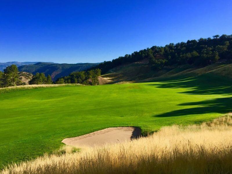 A lush green golf course, perfect for a relaxing round of golf, is surrounded by tall trees and hills under a clear blue sky. A sand bunker is visible in the foreground, with shadows stretching across the vibrant grass.