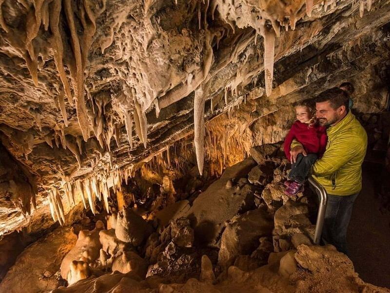 A man in a green jacket holds a child in a cave, surrounded by stalactites and stalagmites. They marvel at the impressive rock formations under warm lighting, enjoying a unique spring break adventure.