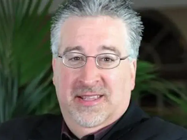 A man with short gray hair, glasses, and a goatee, reminiscent of Larry's distinctive style, is wearing a black suit jacket. The background is blurred with green plant leaves gently visible.