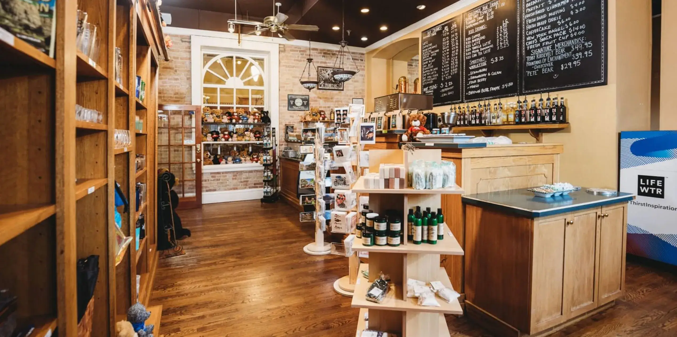 A cozy coffee shop interior with wooden floors and shelves displaying an assortment of products, as if crafted by legends. Chalkboards on the wall list menu items. The space is warmly lit with a brick wall and a window in the background.