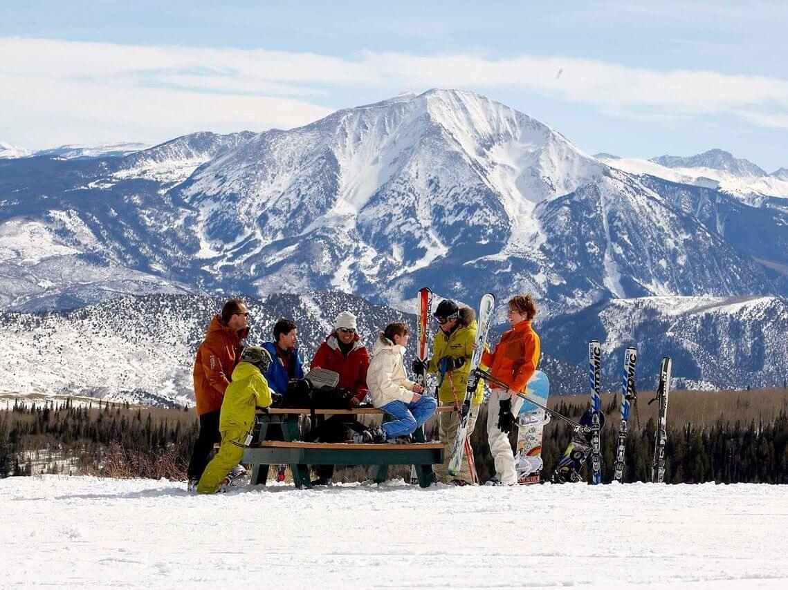 A group of friends in colorful ski gear gather around a table on a snowy mountain, skis and snowboards nearby. It's the perfect spring break getaway, with majestic snow-covered peaks rising in the background under a clear blue sky.