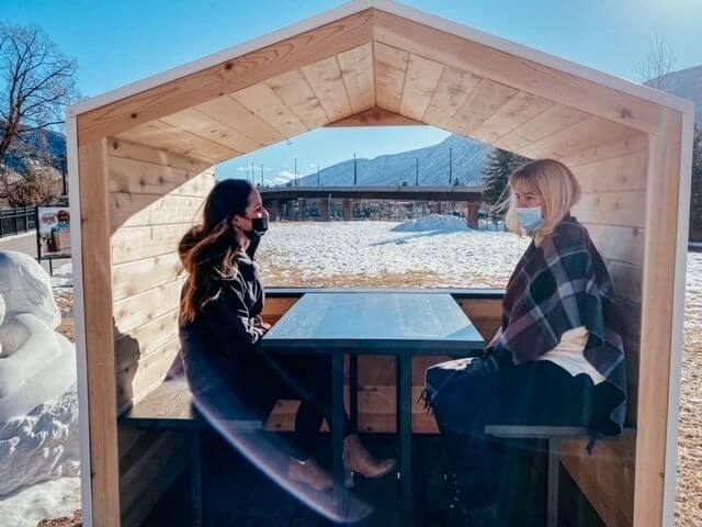 Two people wearing masks sit at a wooden outdoor table under a small shelter. The snow-covered ground enhances the serene beauty of the scene, with mountains providing a breathtaking backdrop under the clear blue sky—a perfect setting for an outdoor escape.