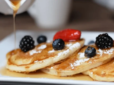 A delightful breakfast awaits: three pancakes topped with blackberries, blueberries, and a strawberry, all drizzled with syrup and dusted with powdered sugar on a white plate. A stream of syrup cascades over them from the left, inviting indulgence.