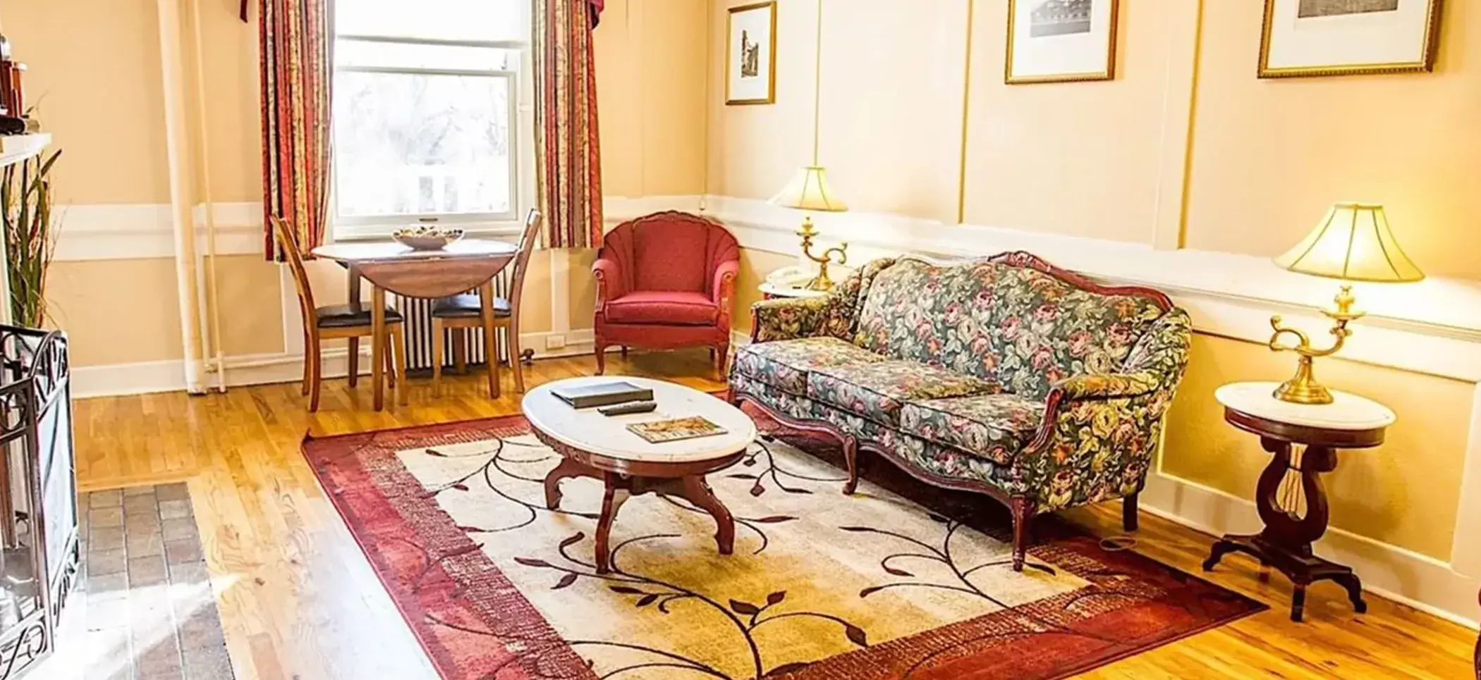 A cozy living room showcases a floral-patterned sofa and red armchair around a wooden coffee table on a patterned rug. Two lamps flank the sofa, embodying its signature charm. Nearby, a small dining table sits by the window, with light walls adorned with framed pictures.