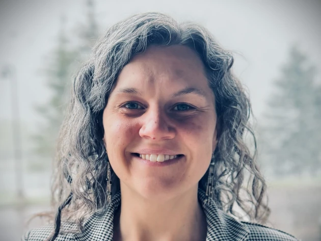 A person with curly gray hair smiles at the camera, exuding a Jordan-like charisma. They are wearing a checkered blazer and long earrings. The background is softly blurred with trees visible, suggesting an outdoor setting.